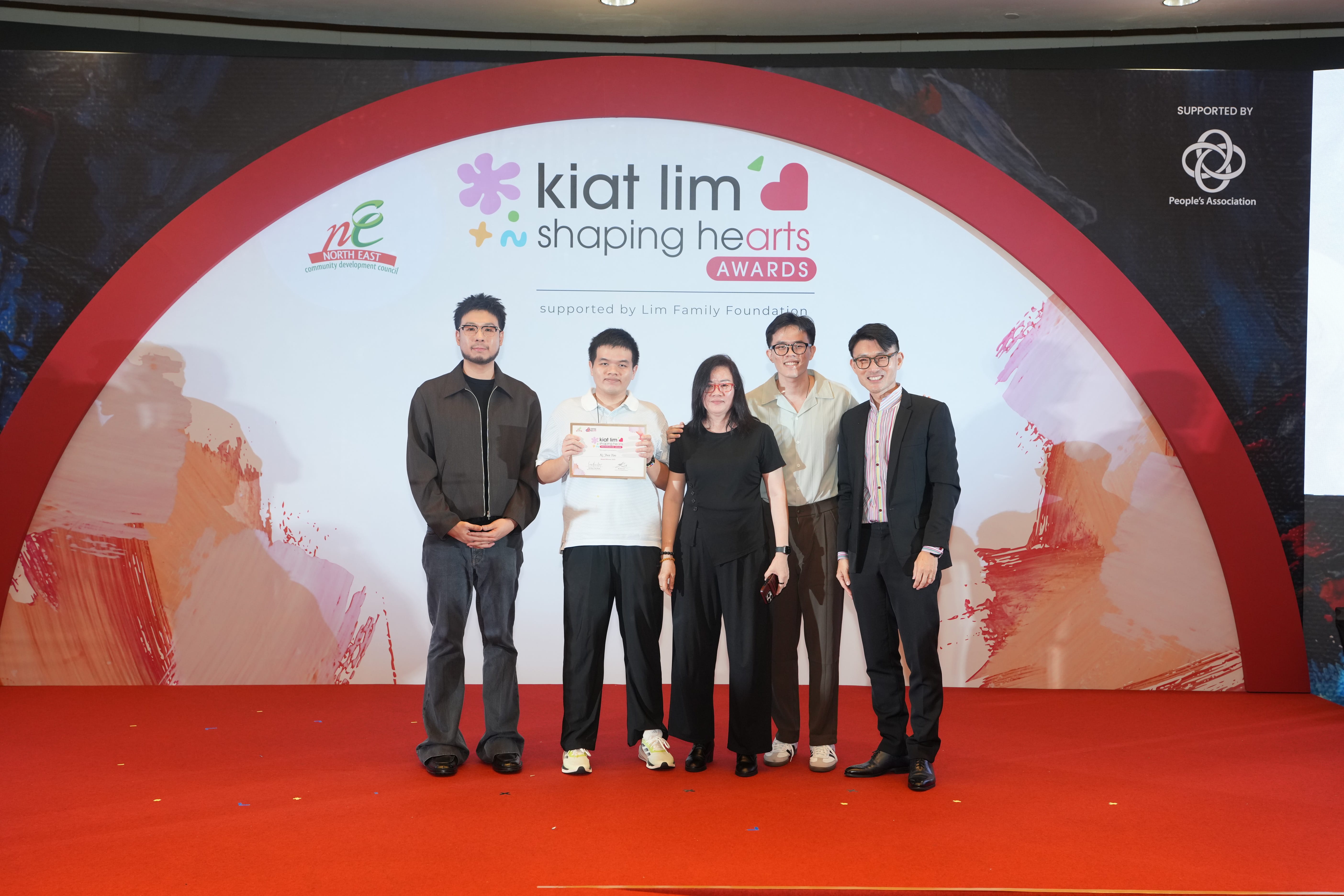 Award recipient standing on stage with the presenter, holding their certificate and posing for a group photo during the award ceremony, with a backdrop reading ‘Kiat Lim-Shaping Hearts Awards'.